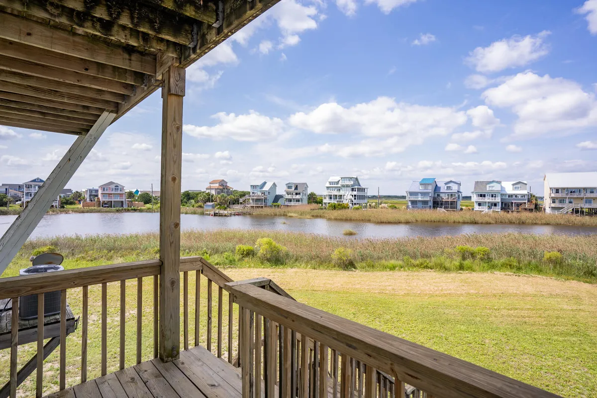 A tranquil waterside scene with houses lining the shore, abundant greenery, and a glassy water surface. The deck area encourages birdwatching, fishing, and relaxing as the sun sets, all within a natural, unspoiled environment.