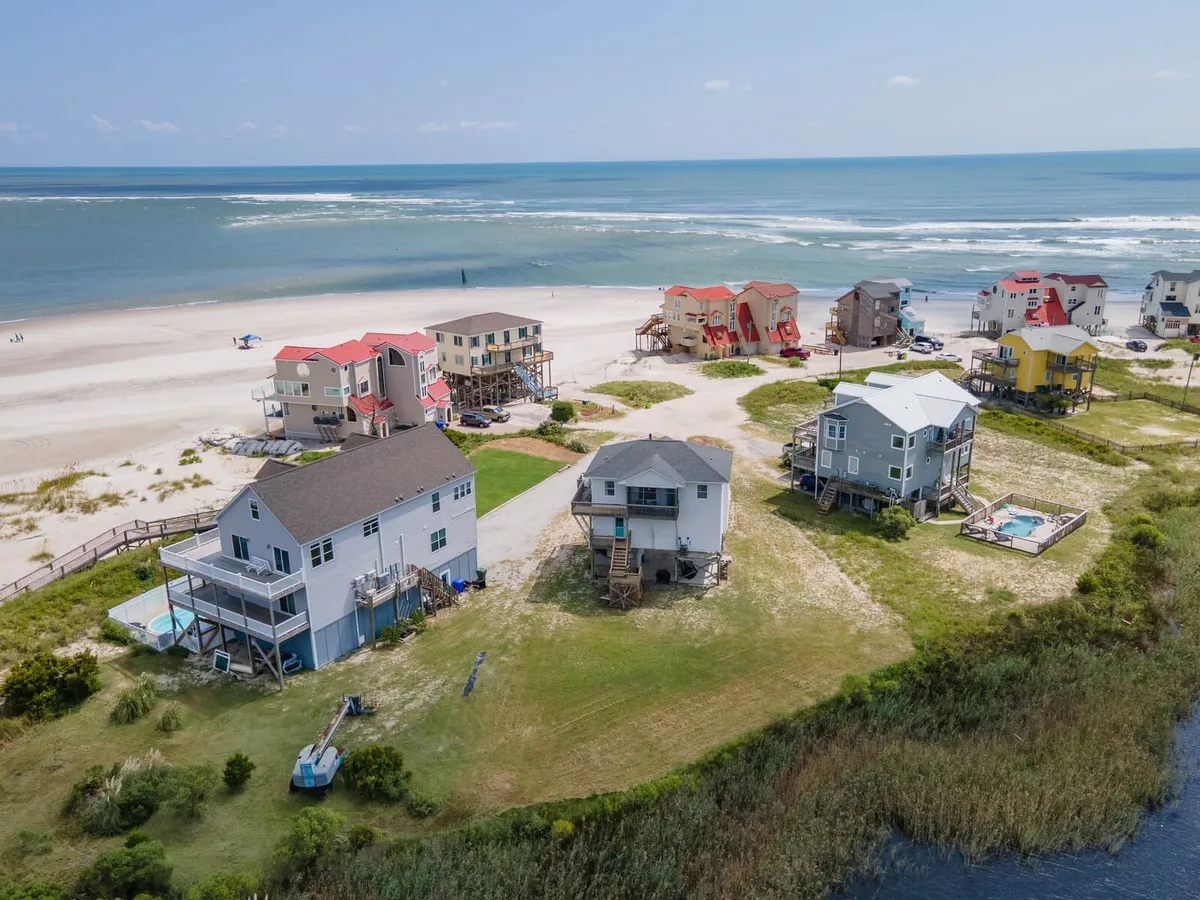 A vibrant exterior scene featuring beachfront houses, a sparkling swimming pool, sand dunes, ocean waves, and lush green vegetation. Families are seen swimming, beachcombing, sunbathing, and gathering for picnics. The view extends to the beach, conveying the close proximity and easy access to water activities.