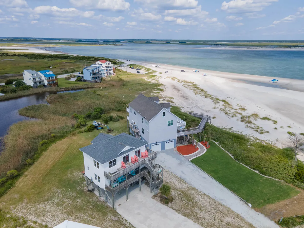 The exterior of a beachfront property with colorful beach chairs on a spacious deck facing the ocean. The scene showcases sandy dunes, ocean waves, and open skies, inviting families to enjoy barbecues, sunsets, and direct access to water activities.