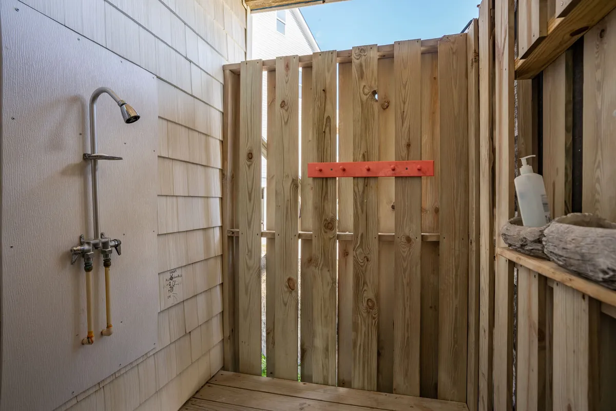 A charming and private outdoor shower is featured, enclosed by a wooden fence and accented with a red detail, offering a rejuvenating space for rinsing off after a day at the beach. The setup is both functional and visually appealing, evoking a sense of refreshment and beachside adventure.