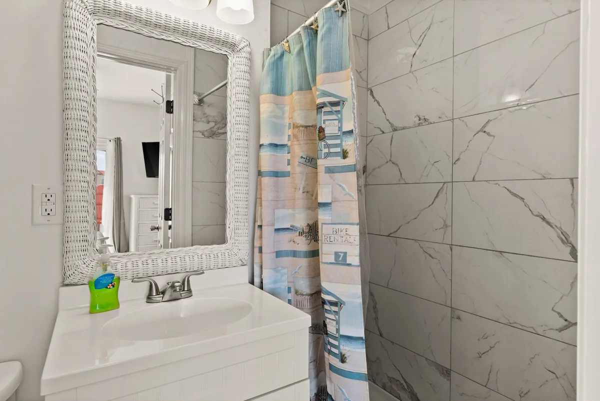 A bright, inviting bathroom featuring a modern sink, marble tiles, a white wicker mirror, and a colorful shower curtain. The space feels fresh and well-lit, ideal for personal grooming and relaxation.