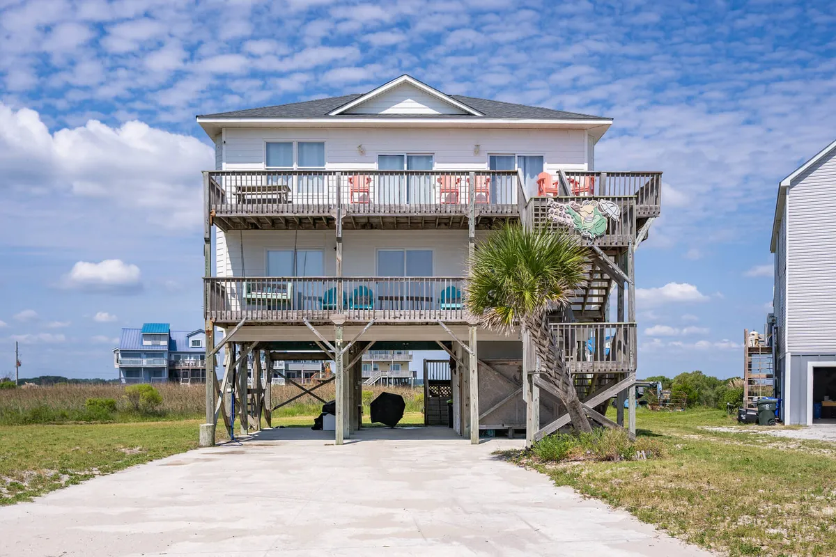 An exterior shot showcasing the property's two balconies framed by a palm tree and a prominent staircase. The balconies are equipped with lounge chairs and offer sweeping views, hinting at both sunrise and sunset vistas.
