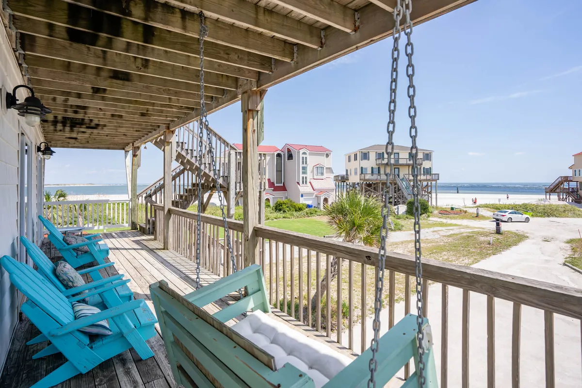 A tranquil deck scene with turquoise rocking chairs facing the beach, overlooking the ocean and surrounded by neighboring houses. The wooden deck provides a welcoming space for socializing, enjoying the view, or sipping morning coffee to the sound of the waves.