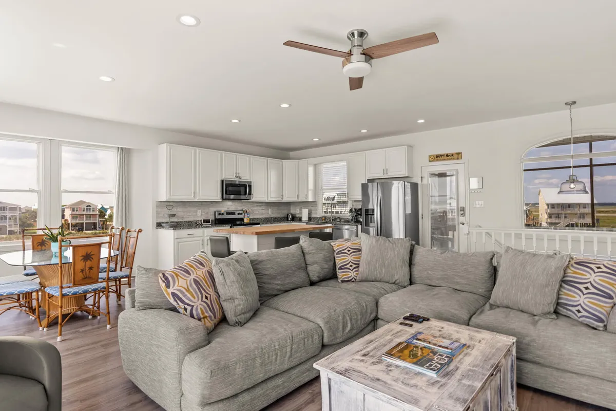 An open-concept great room featuring a spacious sectional sofa, a dining area with wooden chairs, and a modern kitchen with stainless appliances and large windows that fill the space with natural light. The expansive windows offer breathtaking water views, creating a seamless connection between the living, dining, and kitchen areas.