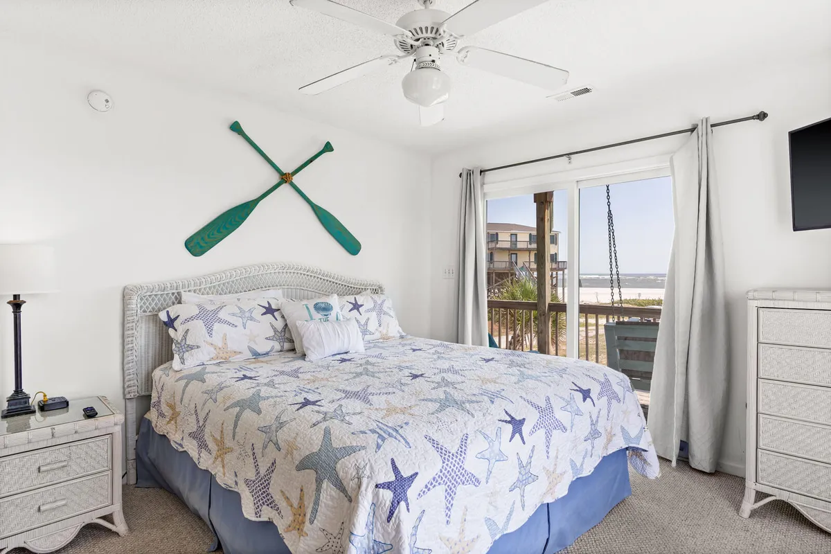 A cozy bedroom with a starfish-patterned quilt, decorative wooden paddles on the wall, and a window that frames a beautiful beach scene. The room’s soft hues and inviting textures create a welcoming, restful environment.