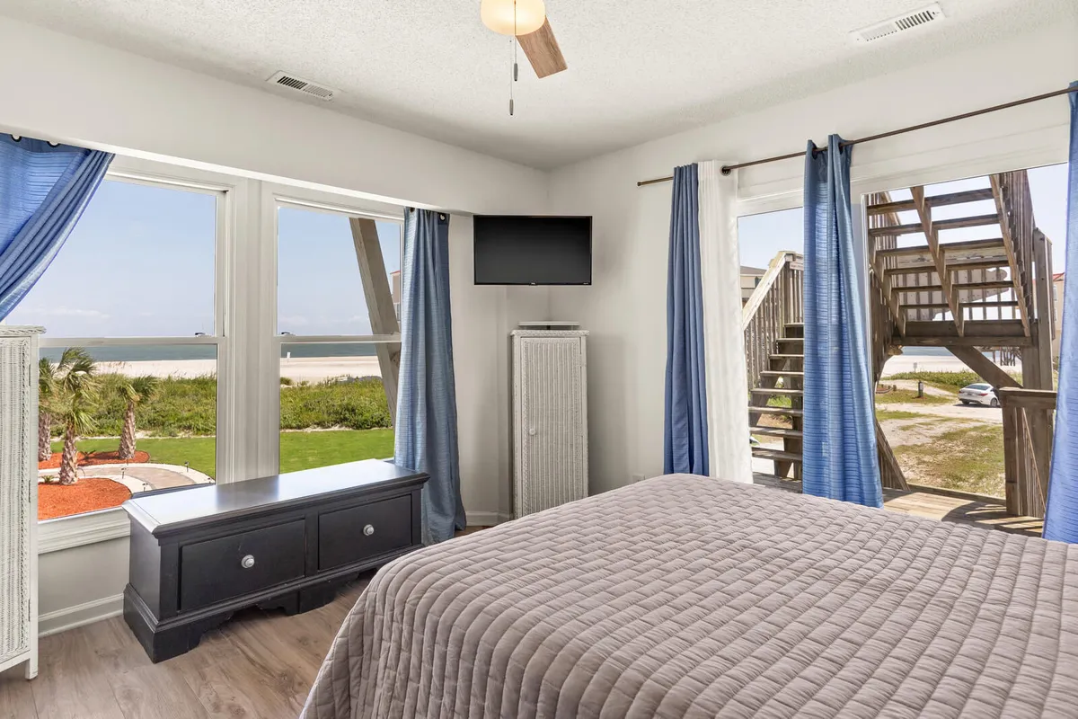 A tranquil bedroom with a large, comfortable bed positioned to face oversized windows that offer an expansive ocean view. The room is styled with serene decor and a stylish dresser that enhances the sense of luxury and peace. Natural light fills the space, highlighting the plush linens and thoughtful design details.