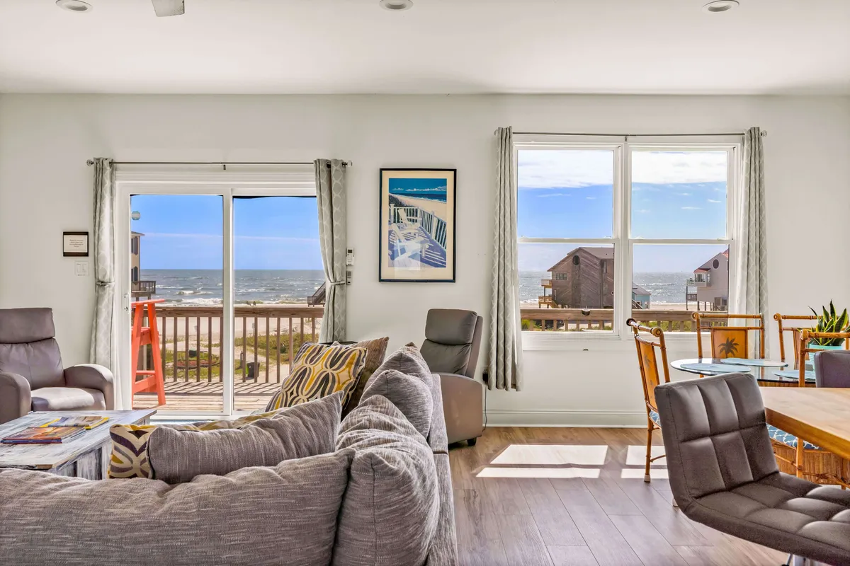 A bright, inviting interior scene with large windows overlooking the ocean, a comfortable seating area, and a dining table set near the view. The furnishings offer a cozy yet elegant environment for meals, reading, or socializing with friends against a backdrop of sea and sky.