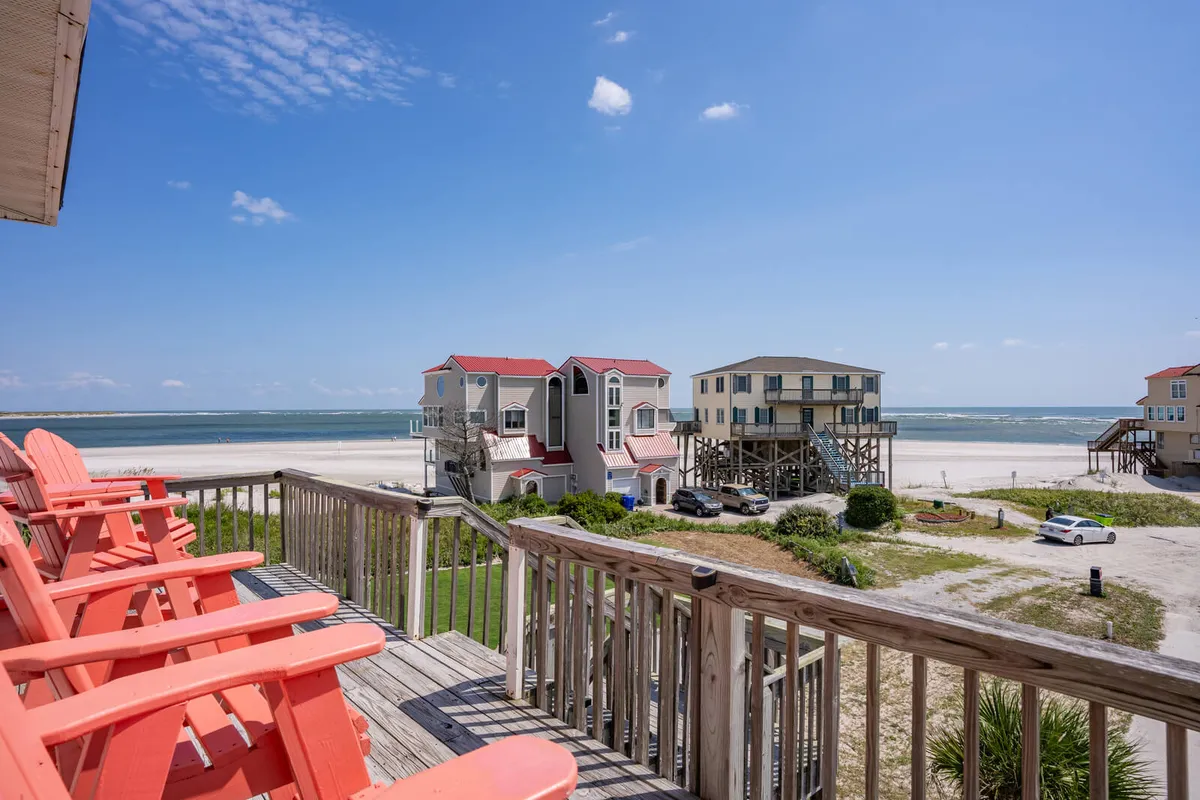 A beachfront scene featuring red Adirondack chairs positioned on a deck with a direct ocean view, flanked by colorful houses. The open, inviting setup encourages relaxation and socializing while taking in the beach and water.