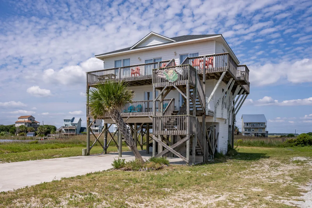 This image captures the exterior of the house with a prominent wooden deck furnished with chairs, an inviting palm tree in the foreground, and the elevated structure hinting at outdoor living spaces ideal for relaxing and dining. The scene is bathed in warm, golden light, evoking the feeling of sunset by the sea.