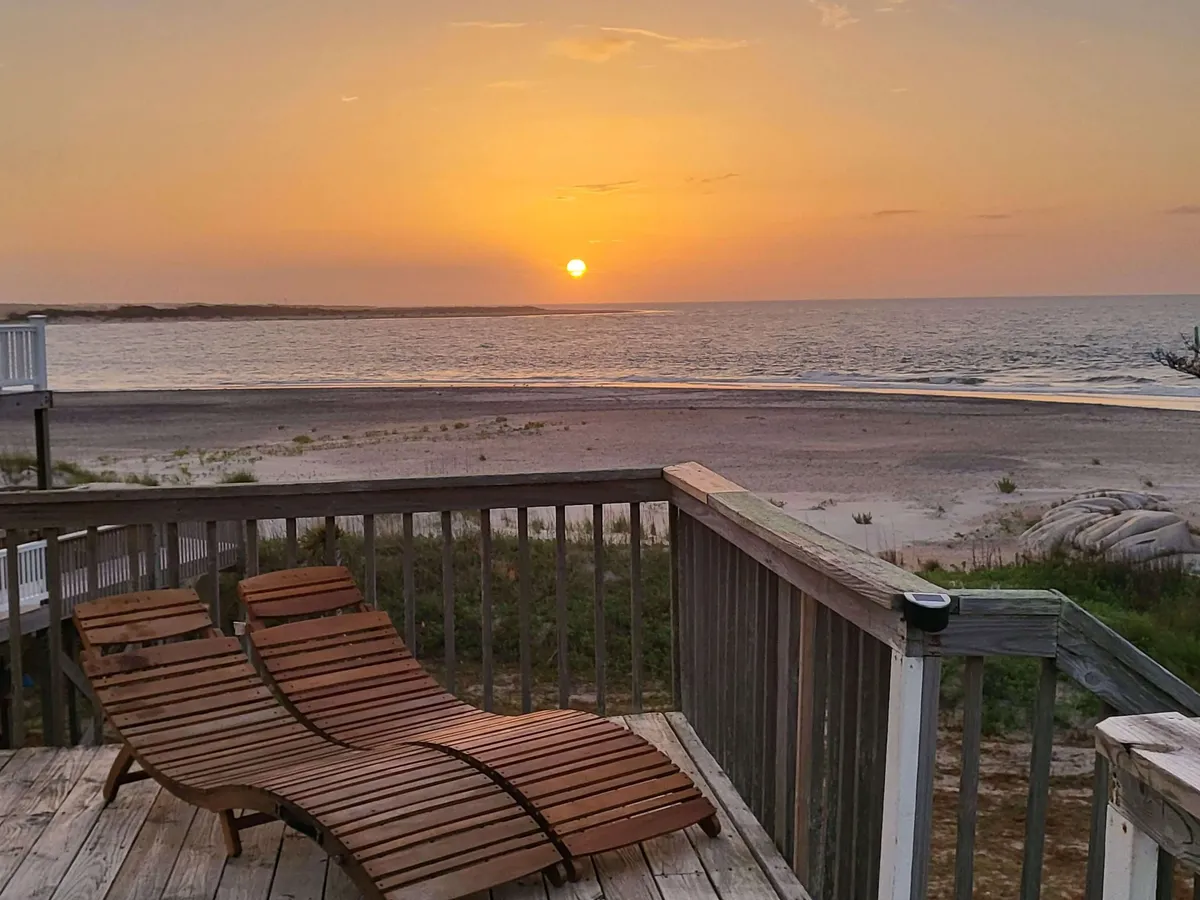 The image showcases a serene sunset over the ocean, viewed from a deck with inviting wooden lounge chairs overlooking an expansive, peaceful beach. The colors of dusk reflect off the water, creating a tranquil and dreamy coastal atmosphere perfect for unwinding.
