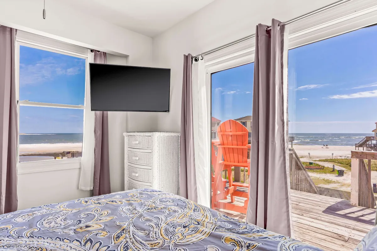 A calming bedroom featuring a patterned bedspread, large windows with a stunning ocean view, and a pop of color from a red outdoor chair. The layout highlights guest comfort with a cozy atmosphere perfect for relaxation and unwinding after sun-filled days.